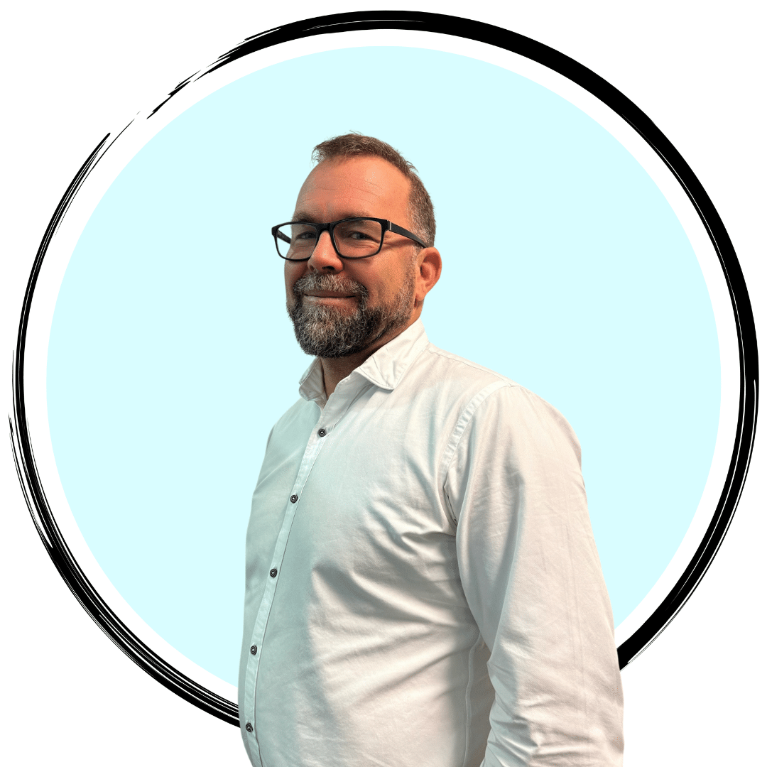 A man with short hair and a beard is wearing glasses and a white button-up shirt. He is standing and looking at the camera with a slight smile. The background is a light blue circle outlined in black.