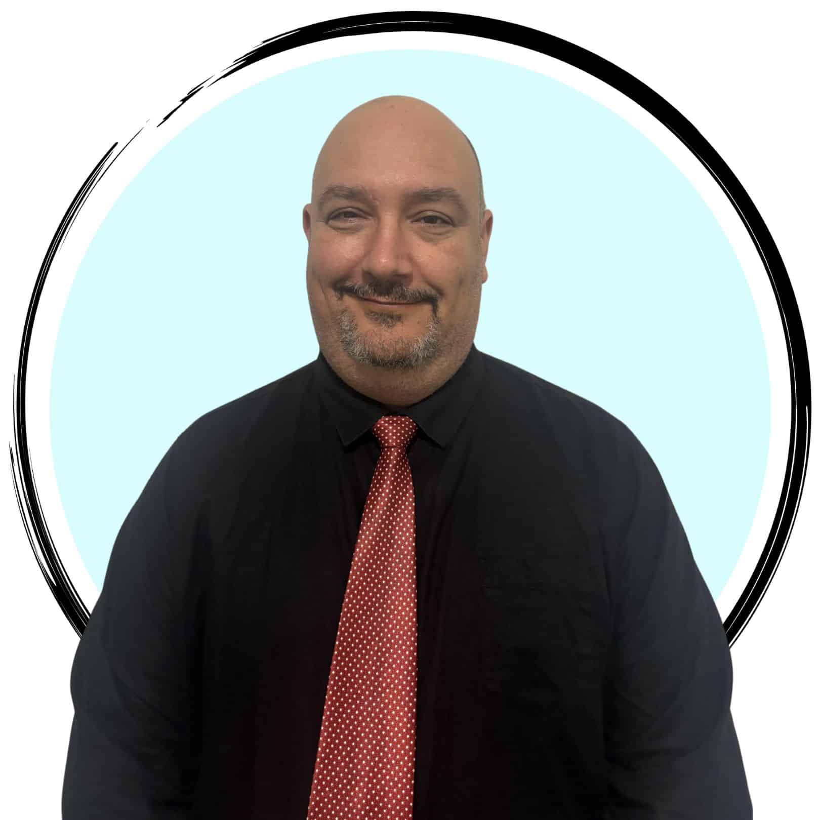 A bald, middle-aged man with a goatee is wearing a black shirt and red tie with white polka dots. He is smiling slightly and stands in front of a light blue circular background with a black outline.